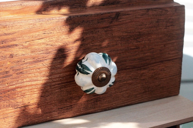 May include: White ceramic drawer knob with a green leaf design. The knob is mounted on a wooden drawer front.