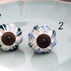 May include: Two white ceramic cabinet knobs with a floral design. One knob has blue flowers and green leaves, the other has green leaves and white flowers. Both knobs have a brown metal base.