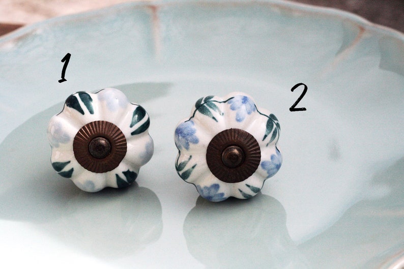 May include: Two white ceramic cabinet knobs with a floral design. One knob has blue flowers and green leaves, the other has green leaves and white flowers. Both knobs have a brown metal base.