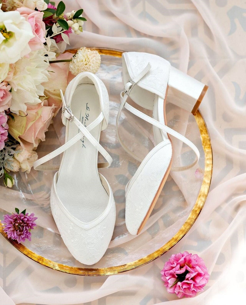 K&ouml;nnte beinhalten: Ein Paar wei&szlig;e Satin-Brautschuhe mit zarten Spitzendetails und einem kleinen Blockabsatz. Die Schuhe befinden sich auf einem goldenen Teller mit einem Blumenstrau&szlig; im Hintergrund.