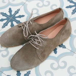 May include: A pair of brown suede lace-up shoes with a rounded toe. The shoes are on a white and blue patterned tile floor.