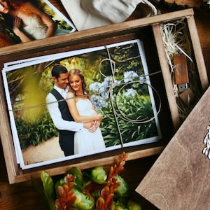 May include: A wooden box with a lid, containing a stack of printed photographs of a couple in a garden setting. The box also contains a USB drive with a wooden casing. The lid of the box has a decorative design of a wreath.