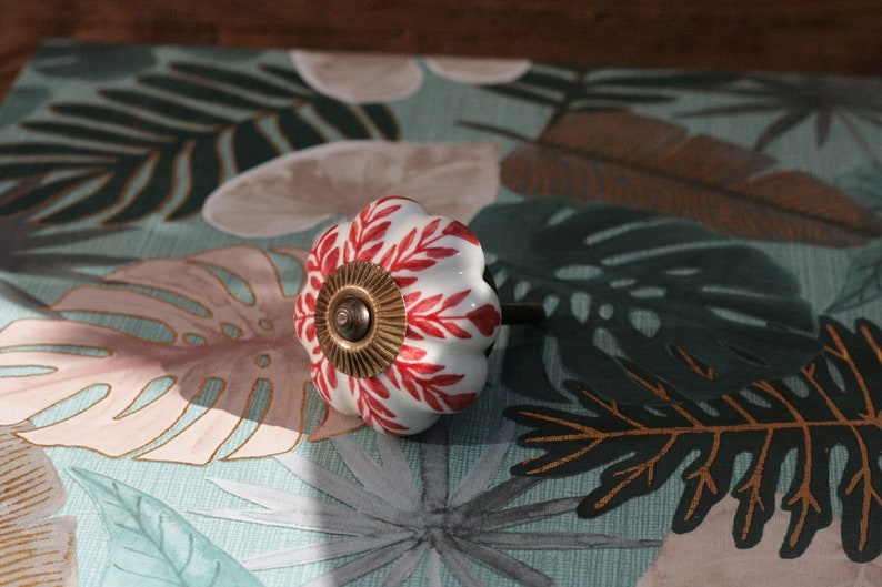 K&ouml;nnte beinhalten: Wei&szlig;er Keramik-Schrankknopf mit rotem und goldenem Blumendesign. Der Knopf ist k&uuml;rbisf&ouml;rmig und hat ein kleines Schraubenloch auf der R&uuml;ckseite.
