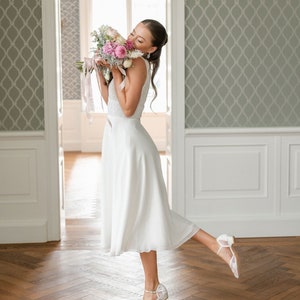 May include: A woman in a white sleeveless dress holds a bouquet of pink and white flowers. She is wearing white lace heels. The background features a light green patterned wallpaper and a wooden floor.