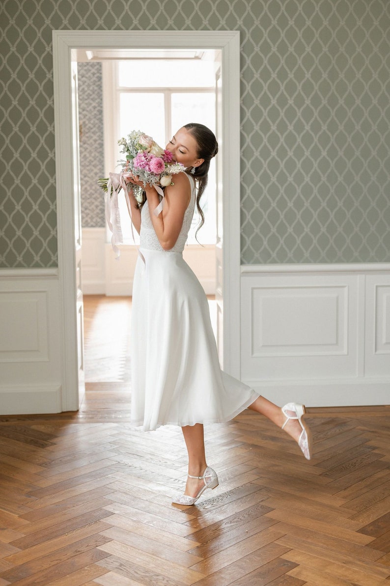 May include: A woman in a white sleeveless dress holds a bouquet of pink and white flowers. She is wearing white lace heels. The background features a light green patterned wallpaper and a wooden floor.