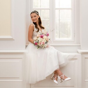 K&ouml;nnte beinhalten: Eine Braut in einem wei&szlig;en Brautkleid mit einem Spitzenoberteil und einem T&uuml;llrock. Sie h&auml;lt einen Strau&szlig; aus rosa und wei&szlig;en Blumen und sitzt auf einer Fensterbank.