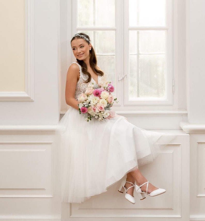 K&ouml;nnte beinhalten: Eine Braut in einem wei&szlig;en Brautkleid mit einem Spitzenoberteil und einem T&uuml;llrock. Sie h&auml;lt einen Strau&szlig; aus rosa und wei&szlig;en Blumen und sitzt auf einer Fensterbank.