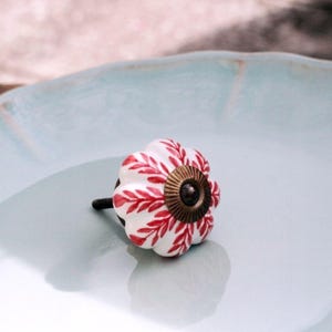 May include: A white ceramic cabinet knob with a scalloped, flower-like shape featuring a red leaf pattern. The center of the knob has a circular, ribbed bronze-toned metal plate with a dark screw head in the middle and a black metal bolt extending.