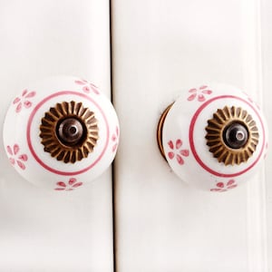 Könnte beinhalten: Zwei weiße Keramik-Schrankknöpfe mit rosafarbenen Blumendesigns und goldenen Akzenten. Die Knöpfe haben ein erhabenes, kreisförmiges Design in der Mitte.