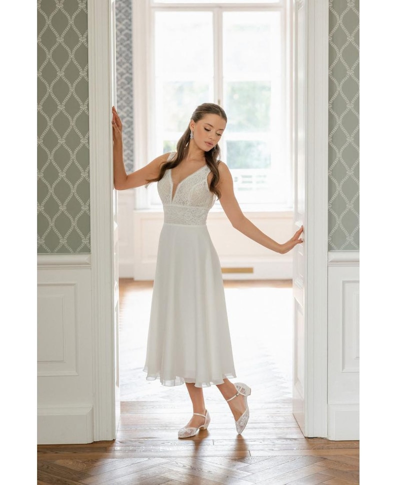 May include: A white wedding dress with a V-neckline and lace bodice. The dress has a flowing skirt that falls to the mid-calf. The bride is wearing white lace shoes with a small heel. The setting is a bright room with white doors and a window.