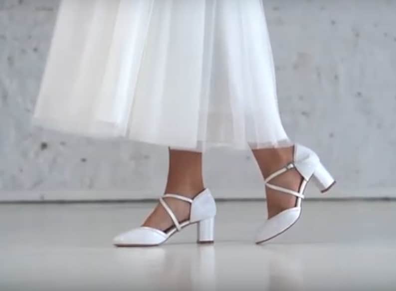 K&ouml;nnte beinhalten: Wei&szlig;e Brautschuhe mit einem Kreuzriemen-Design und einem Blockabsatz.