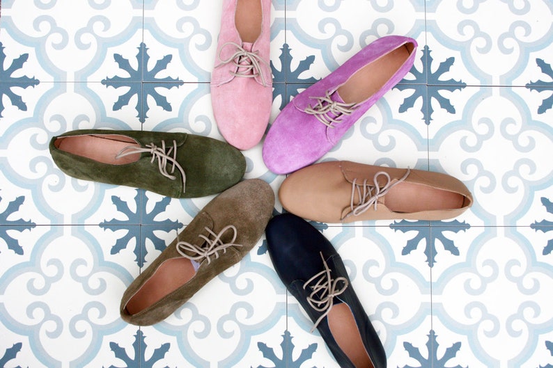 May include: Six pairs of suede lace-up shoes in different colors, including pink, purple, green, brown, beige, and blue, arranged in a star shape on a white and blue tiled floor.