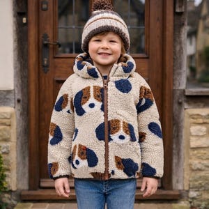 Könnte beinhalten: Eine beigefarbene Kapuzenjacke aus Sherpa-Fleece mit einem sich wiederholenden Muster aus braunen, weißen und marineblauen Hundegesichtern, kombiniert mit einer passenden Strickmütze mit braunem Bommel und einer blauen Jeans.