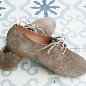 May include: A pair of brown suede lace-up shoes with white laces. The shoes are on a tiled floor with a blue and white pattern.
