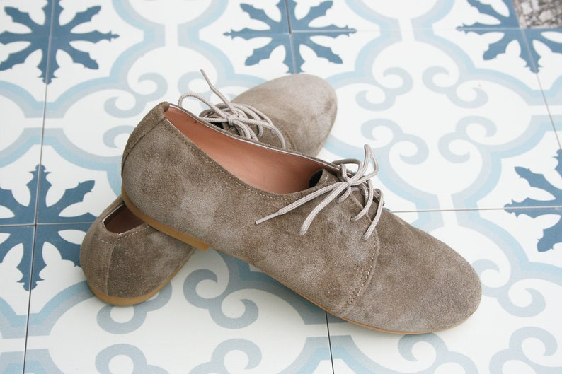 May include: A pair of brown suede lace-up shoes with white laces. The shoes are on a tiled floor with a blue and white pattern.