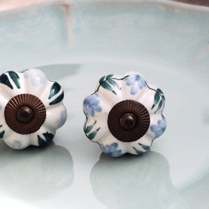 May include: Two white ceramic cabinet knobs with a floral design. The knobs have a blue and green floral pattern and a brass center.