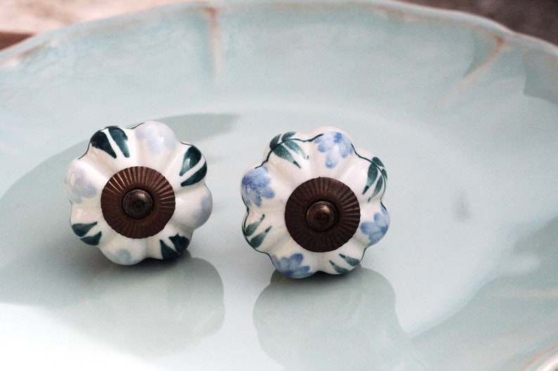 May include: Two white ceramic cabinet knobs with a floral design. The knobs have a blue and green floral pattern and a brass center.