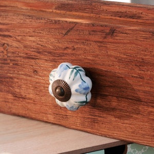 May include: A white ceramic drawer knob with a blue and green floral design. The knob is attached to a wooden drawer.