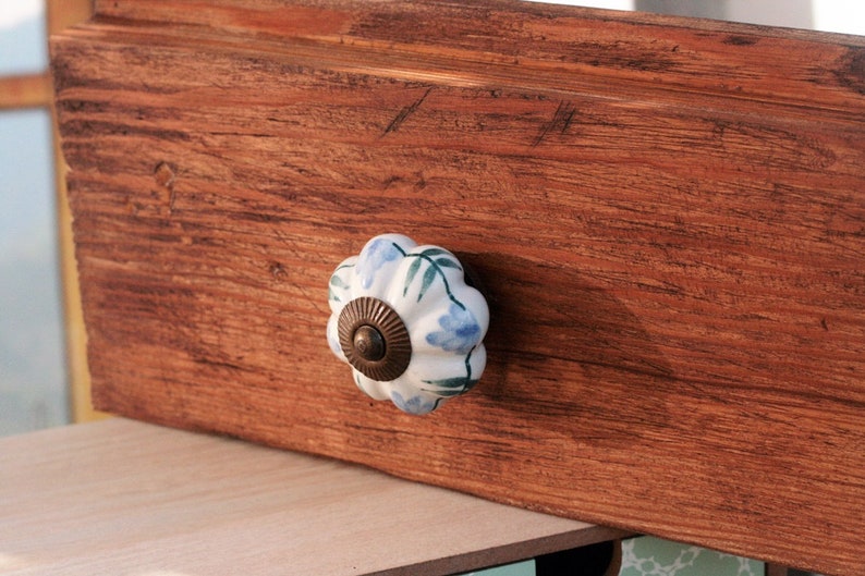 May include: A white ceramic drawer knob with a blue and green floral design. The knob is attached to a wooden drawer.