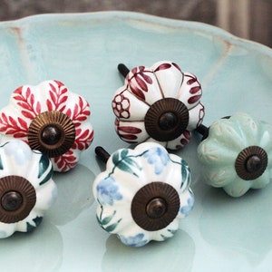 May include: Five ceramic cabinet knobs with different floral patterns. The knobs are white with red, blue, green, and light green floral designs. Each knob has a brass-colored base.