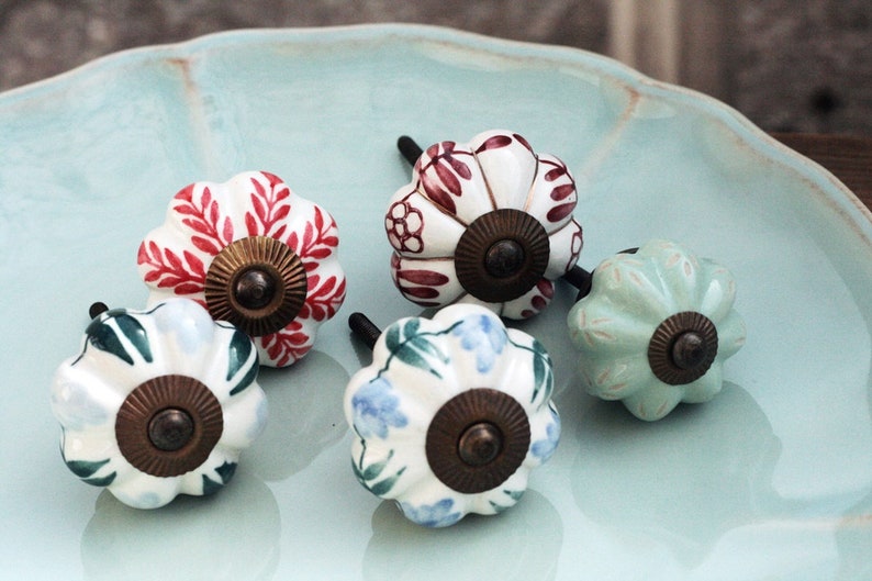 May include: Five ceramic cabinet knobs with different floral patterns. The knobs are white with red, blue, green, and light green floral designs. Each knob has a brass-colored base.