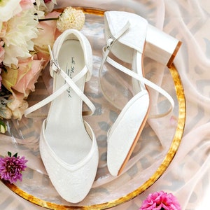 K&ouml;nnte beinhalten: Ein Paar wei&szlig;e Braut-Schuhe aus Spitze mit Kn&ouml;chelriemen und Abs&auml;tzen. Die Schuhe befinden sich auf einem goldenen und wei&szlig;en Teller mit Blumen.