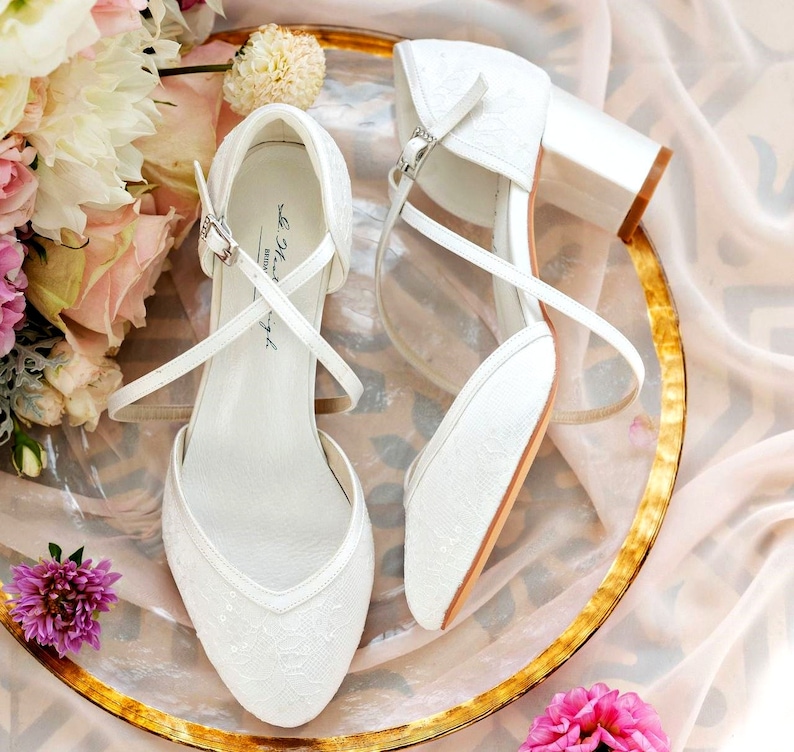 K&ouml;nnte beinhalten: Ein Paar wei&szlig;e Braut-Schuhe aus Spitze mit Kn&ouml;chelriemen und Abs&auml;tzen. Die Schuhe befinden sich auf einem goldenen und wei&szlig;en Teller mit Blumen.