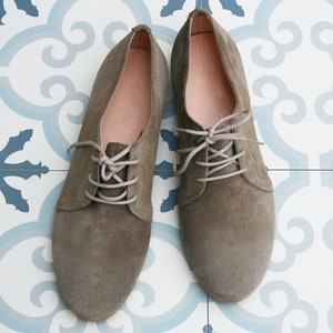 May include: A pair of gray suede lace-up shoes with light gray laces. The shoes are on a white and blue patterned tile floor.