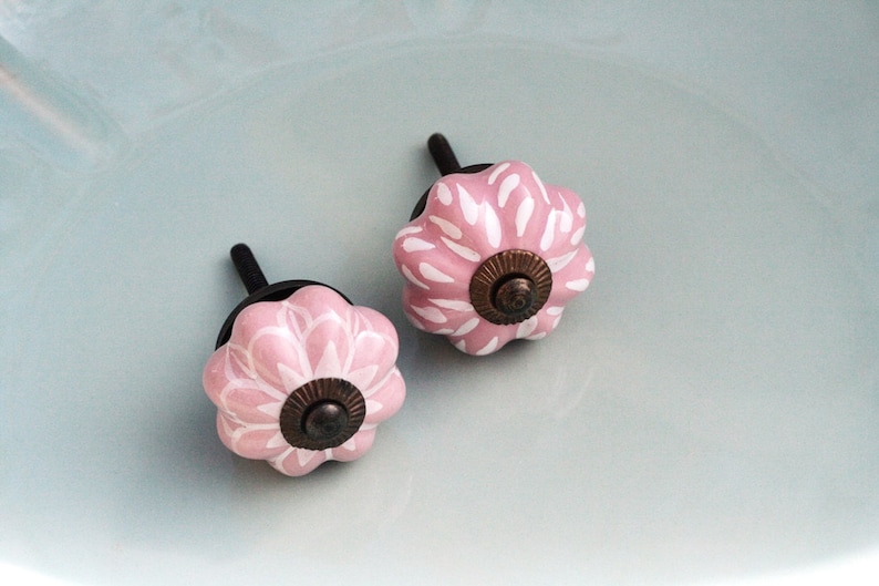 Puede incluir: Dos pomos de caj&oacute;n de cer&aacute;mica rosa con un dise&ntilde;o floral blanco y un centro de bronce. Los pomos tienen un v&aacute;stago de metal negro.