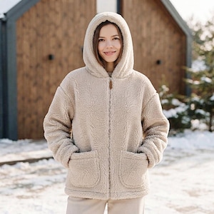 Könnte beinhalten: Eine beigefarbene, langärmelige Kapuzenjacke mit Reißverschluss aus strukturiertem, flauschigem Fleece-Material. Die Jacke verfügt über einen durchgehenden Frontreißverschluss mit braunem Zipper, eine Kapuze und zwei große, quadratische aufgesetzte Taschen.