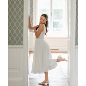 May include: A white sleeveless dress with a lace bodice and a flowing skirt. The dress is paired with white, low-heeled sandals. The image is taken indoors, with a light-filled background and a decorative wall.