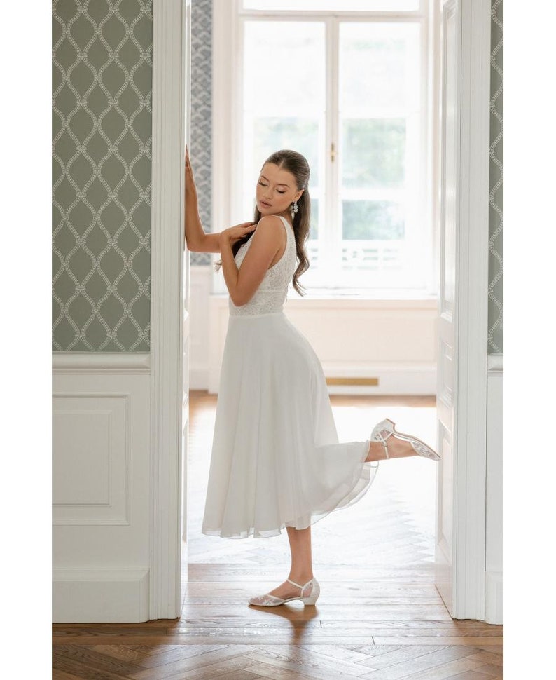 May include: A white sleeveless dress with a lace bodice and a flowing skirt. The dress is paired with white, low-heeled sandals. The image is taken indoors, with a light-filled background and a decorative wall.