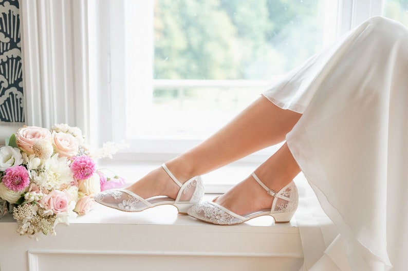 May include: White lace wedding shoes with ankle straps and low heels. The shoes are decorated with floral lace patterns. A bouquet of pink and white flowers sits nearby. The scene is set by a window.