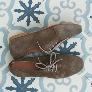 May include: A pair of brown suede lace-up shoes with a light brown sole. The shoes are on a white and blue tiled floor.