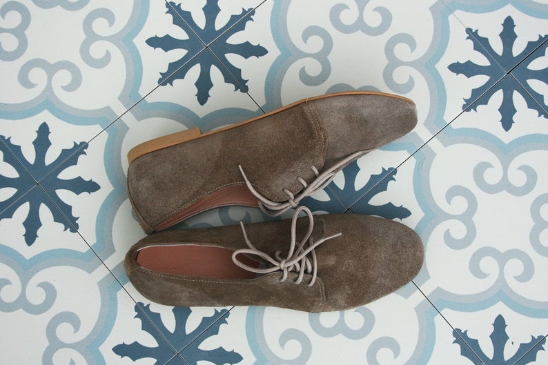 May include: A pair of brown suede lace-up shoes with a light brown sole. The shoes are on a white and blue tiled floor.
