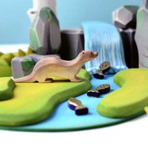 Peut inclure: Une figurine de loutre en bois se tient sur une surface herbeuse verte avec une rivière bleue et une cascade en arrière-plan. La loutre regarde un petit poisson en bois nageant dans la rivière.