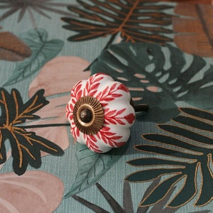 K&ouml;nnte beinhalten: Wei&szlig;er Keramik-Schrankknopf mit rotem und goldenem Blumendesign. Der Knopf ist k&uuml;rbisf&ouml;rmig und hat eine goldene Schraube.