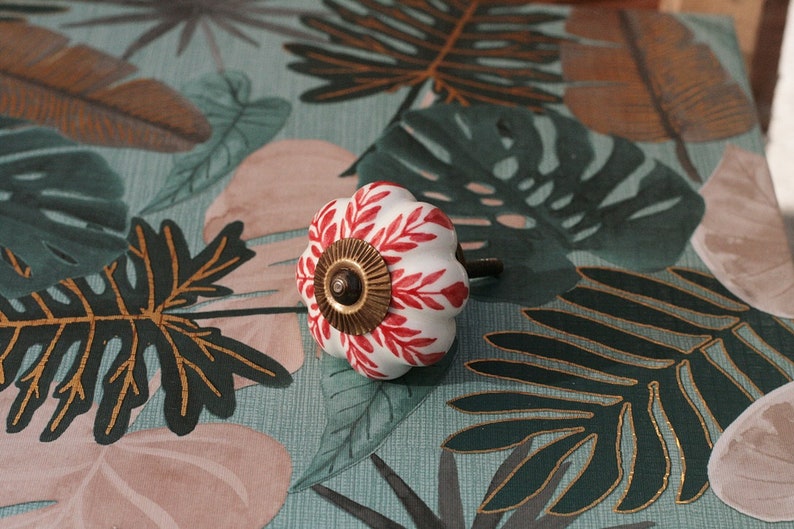 K&ouml;nnte beinhalten: Wei&szlig;er Keramik-Schrankknopf mit rotem und goldenem Blumendesign. Der Knopf ist k&uuml;rbisf&ouml;rmig und hat eine goldene Schraube.