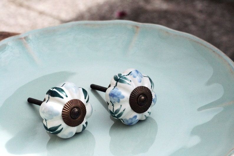 May include: Two white ceramic cabinet knobs with blue and green floral designs and brass accents. The knobs are on a light blue plate.