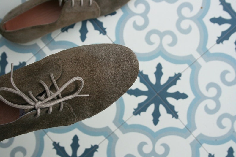 May include: A pair of brown suede lace-up shoes with white laces. The shoes are on a white and blue tiled floor.