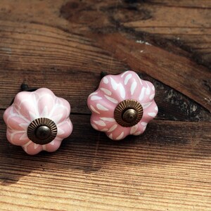 Puede incluir: Dos pomos de armario de cer&aacute;mica rosa con dise&ntilde;os florales blancos y un centro de lat&oacute;n. Los pomos est&aacute;n sobre una superficie de madera.