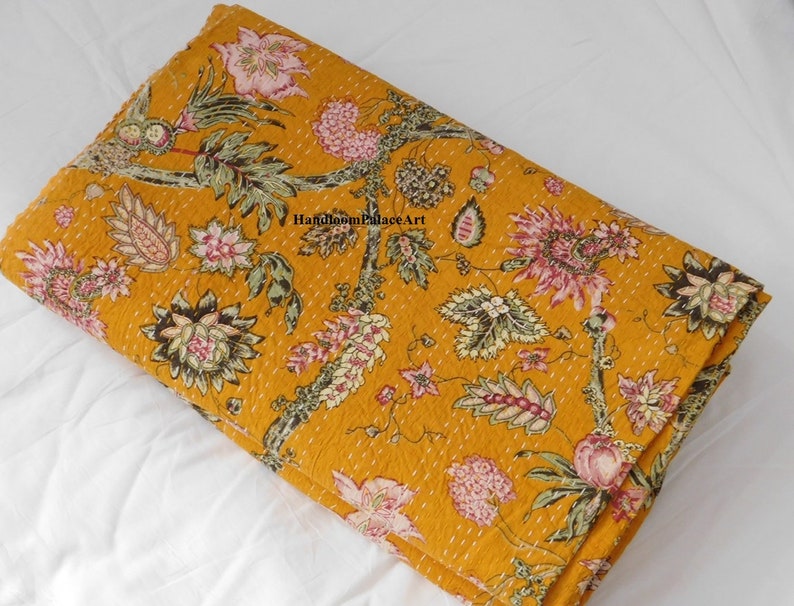 May include: A mustard yellow kantha quilt with a floral pattern. The quilt is made of cotton and has a hand-stitched design. The quilt is folded in half and is lying on a white surface. The text "Handloom Palace Art" is visible in the image.