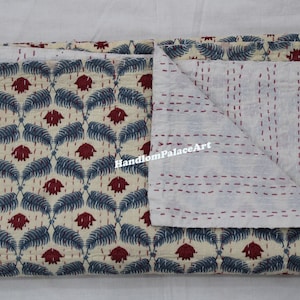 Puede incluir: Una colcha kantha de color crema con un patr&oacute;n floral azul y rojo. La colcha est&aacute; doblada por la mitad, revelando el reverso de algod&oacute;n blanco con costuras rojas.