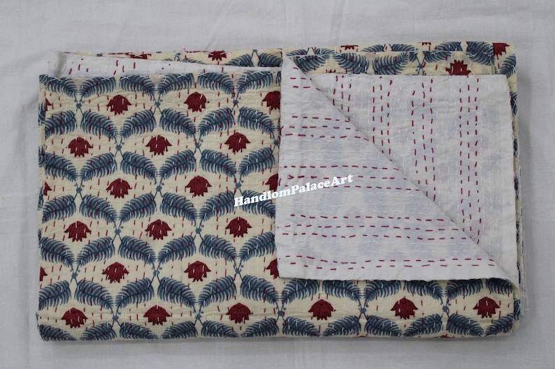 Puede incluir: Una colcha kantha de color crema con un patr&oacute;n floral azul y rojo. La colcha est&aacute; doblada por la mitad, revelando el reverso de algod&oacute;n blanco con costuras rojas.