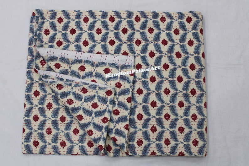 Puede incluir: Una colcha kantha doblada con fondo blanco y un patr&oacute;n repetitivo de hojas azules y flores rojas. La colcha est&aacute; hecha de algod&oacute;n y est&aacute; cosida a mano. El texto "HandloomPalaceArt" es visible en la colcha.