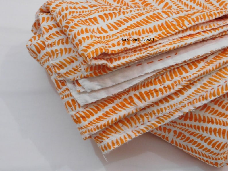 K&ouml;nnte beinhalten: Eine wei&szlig;e und orangefarbene Kantha-Steppdecke mit einem sich wiederholenden Muster aus orangefarbenen Bl&auml;ttern. Die Steppdecke ist dreifach gefaltet und der Stoff ist weich und strukturiert.