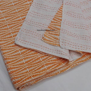 K&ouml;nnte beinhalten: Orange-wei&szlig;e Kantha-Steppdecke mit geometrischem Muster. Die Decke ist in der H&auml;lfte gefaltet und tr&auml;gt ein Etikett mit der Aufschrift "Handloom Palace Art".