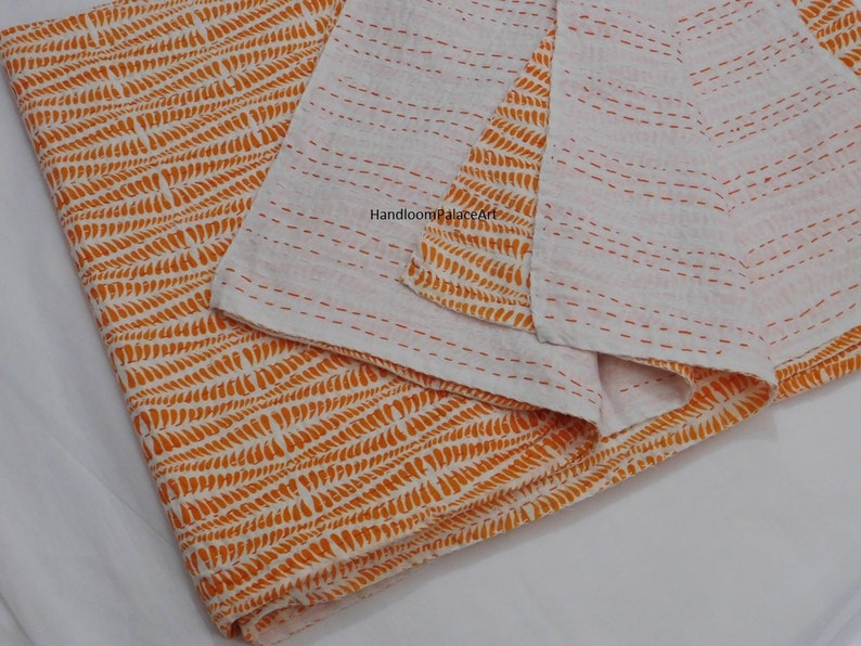 K&ouml;nnte beinhalten: Orange-wei&szlig;e Kantha-Steppdecke mit geometrischem Muster. Die Decke ist in der H&auml;lfte gefaltet und tr&auml;gt ein Etikett mit der Aufschrift "Handloom Palace Art".