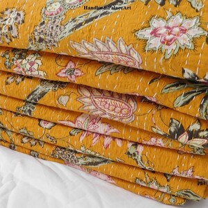 May include: A stack of mustard yellow kantha quilts with a floral print. The quilts are folded and stacked on a white surface. The fabric has a hand-stitched pattern.