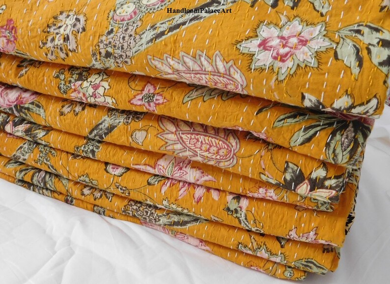 May include: A stack of mustard yellow kantha quilts with a floral print. The quilts are folded and stacked on a white surface. The fabric has a hand-stitched pattern.